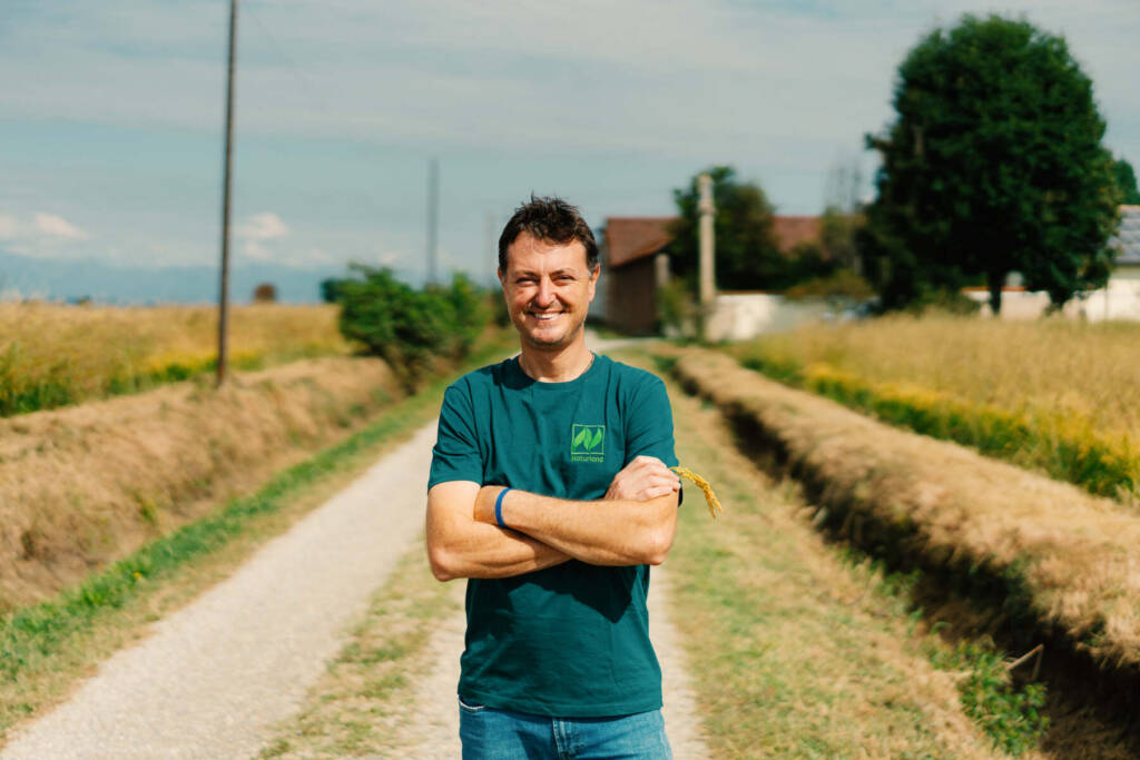 Lächelnder Landwirt steht mit verschränkten Armen auf einem Feldweg, umgeben von Wiesen und einem Bauernhof im Hintergrund.