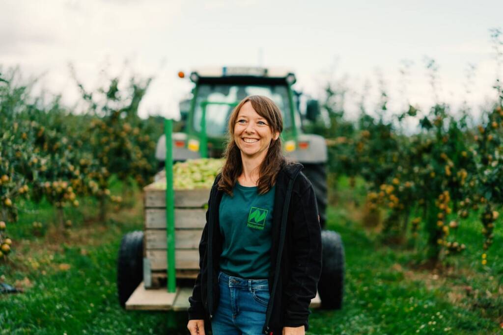 Lächelnde Obstbäuerin steht im Apfelgarten vor einem Traktor mit Kisten voller Äpfel während der Ernte.