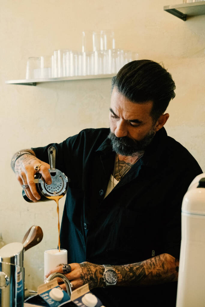 Barista mit Bart gießt frisch zubereiteten Espresso durch ein Sieb in ein Glas in einem Café.